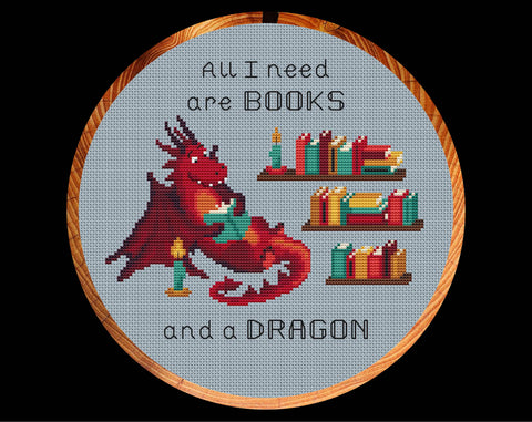 Book Dragon cross stitch pattern. A dragon reading a book next to some bookshelves, with the words 'All I need are BOOKS and a DRAGON'. Shown in hoop.