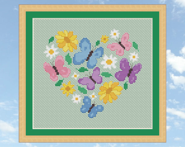 Butterfly Heart cross stitch pattern. Butterflies in pink, purple and blue pastel colours, daisies, yellow flowers and leaves form a heart shape. Shown in frame.