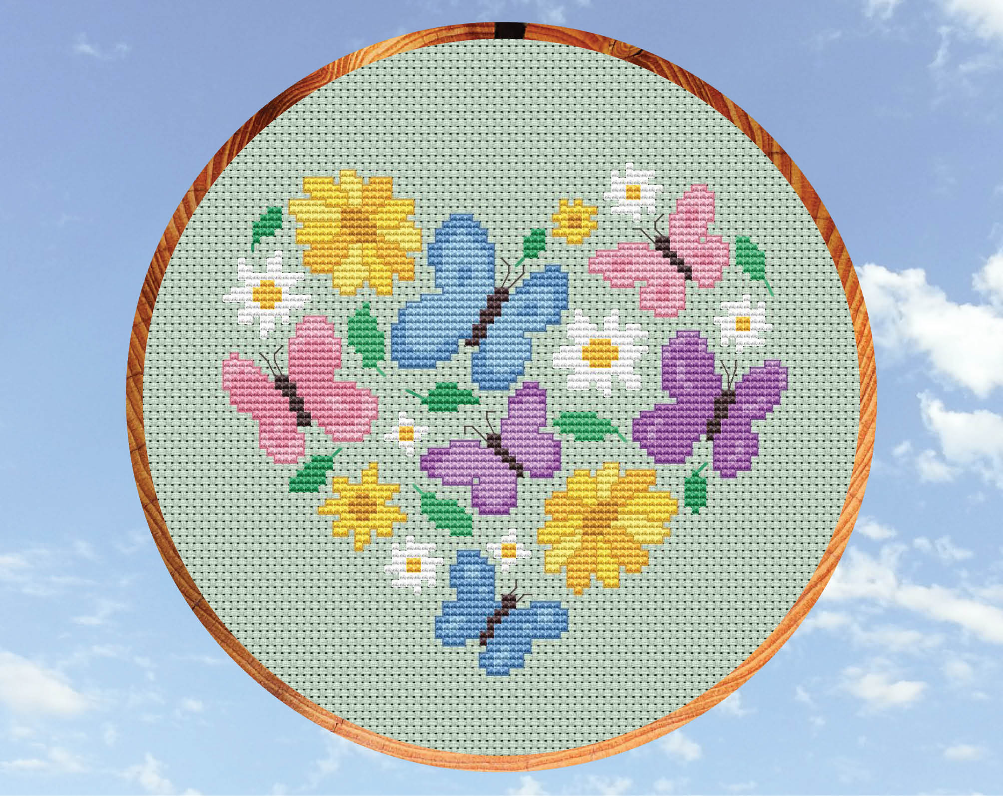 Butterfly Heart cross stitch pattern. Butterflies in pink, purple and blue pastel colours, daisies, yellow flowers and leaves form a heart shape. Shown in hoop.