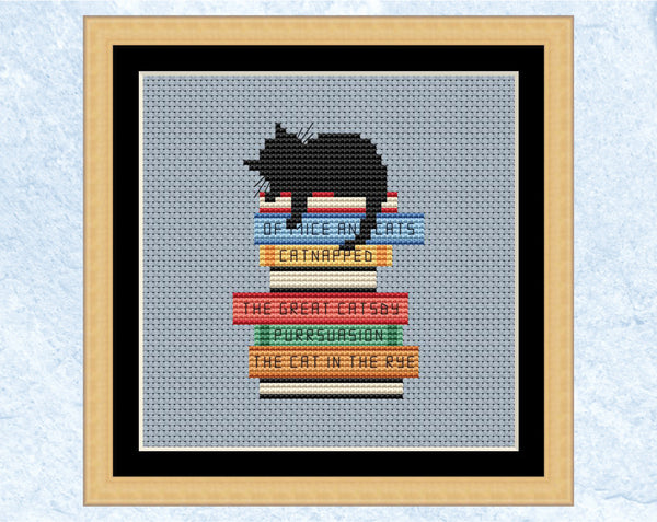 Catnapped cross stitch pattern. Black cat asleep on a pile of books with pun titles. Shown in frame.
