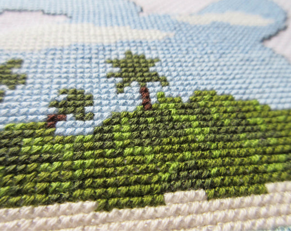 Cross stitch pattern of the silhouette of a turtle filled with a view of a desert island beach, blue sea and sky. Close up view of stitched piece.