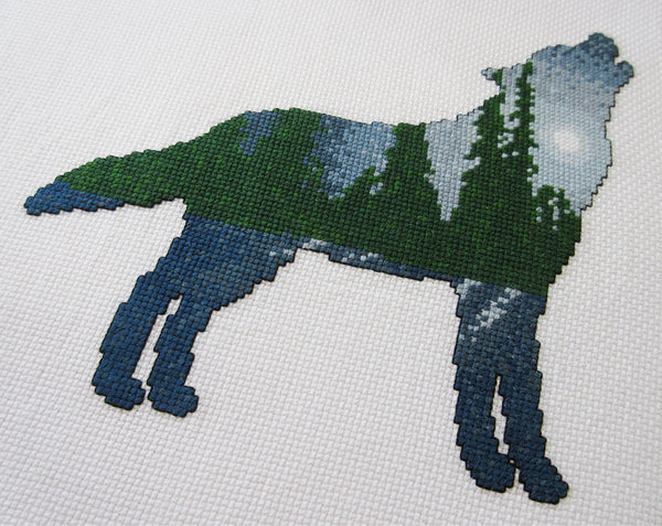 Cross stitch pattern of a wolf silhouette filled with a scene of a moonlit pine forest, with the moonlight reflecting off snow-covered ground. Angled view of stitched piece.