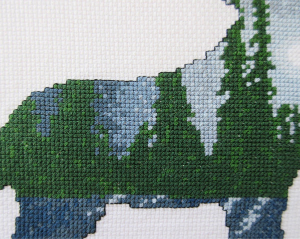 Cross stitch pattern of a wolf silhouette filled with a scene of a moonlit pine forest, with the moonlight reflecting off snow-covered ground. Close up of stitched piece.