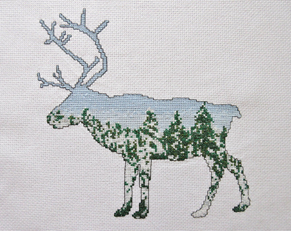 Cross stitch pattern of a reindeer filled with a scene of snow-laden Arctic pine trees against a clear blue sky. Straight view of stitched piece.