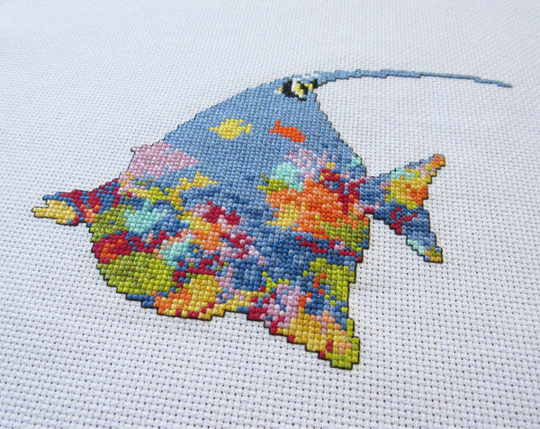 Cross stitch pattern of the silhouette of a tropical fish (a moorish idol fish) filled with a scene of a coral reef. Angled view of stitched piece.