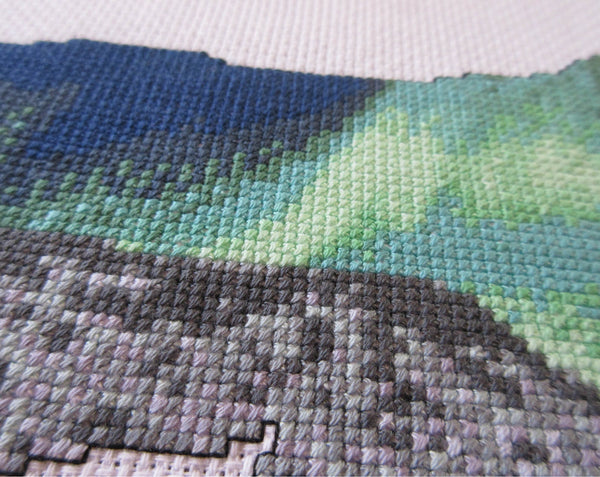 Cross stitch pattern of the silhouette of a polar bear, filled with a scene of the Northern Lights over an Arctic landscape of snow and mountains. Close up angled view of stitched piece.