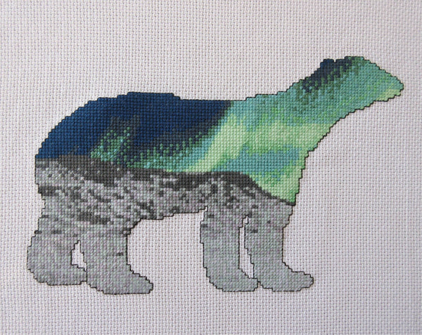 Cross stitch pattern of the silhouette of a polar bear, filled with a scene of the Northern Lights over an Arctic landscape of snow and mountains. Straight view of stitched piece.