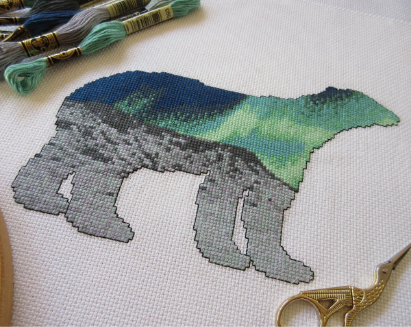 Cross stitch pattern of the silhouette of a polar bear, filled with a scene of the Northern Lights over an Arctic landscape of snow and mountains. Angled view of stitched piece with props.