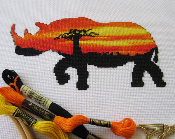 Cross stitch pattern of the silhouette of a rhinoceros filled with a view of an African sunset behind a baobab tree. Angled view of stitched piece and DMC threads.