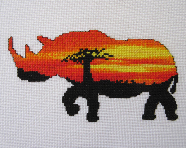 Cross stitch pattern of the silhouette of a rhinoceros filled with a view of an African sunset behind a baobab tree. Straight view of stitched piece.
