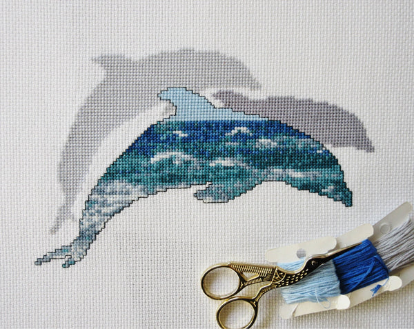 Cross stitch pattern of the silhouette of a dolphin filled with a scene of blue and turquoise ocean waves, with two plain silhouetted dolphins behind it. Stitched piece with scissors and threads.