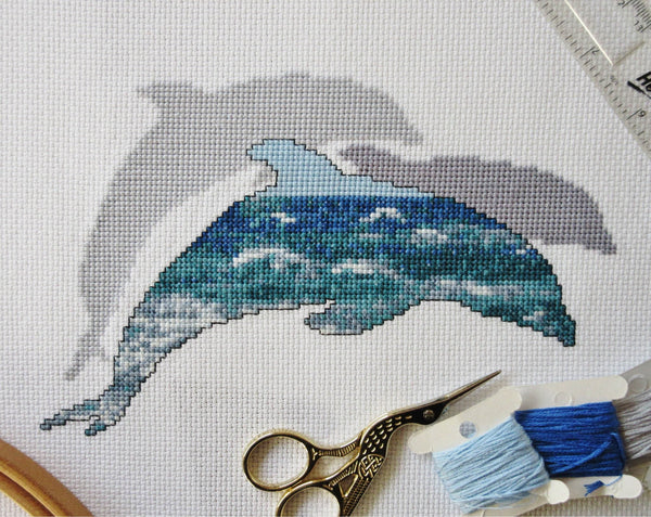 Cross stitch pattern of the silhouette of a dolphin filled with a scene of blue and turquoise ocean waves, with two plain silhouetted dolphins behind it. Stitched piece with props.