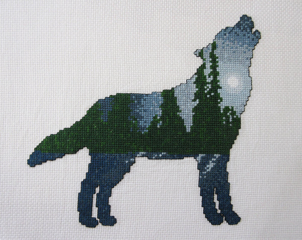 Cross stitch pattern of a wolf silhouette filled with a scene of a moonlit pine forest, with the moonlight reflecting off snow-covered ground. Stitched piece.