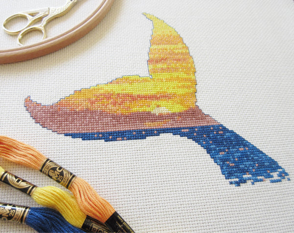 Cross stitch pattern of the silhouette of a whale's tail filled with a scene of a sunset over the ocean. Angled view of stitched piece with props.