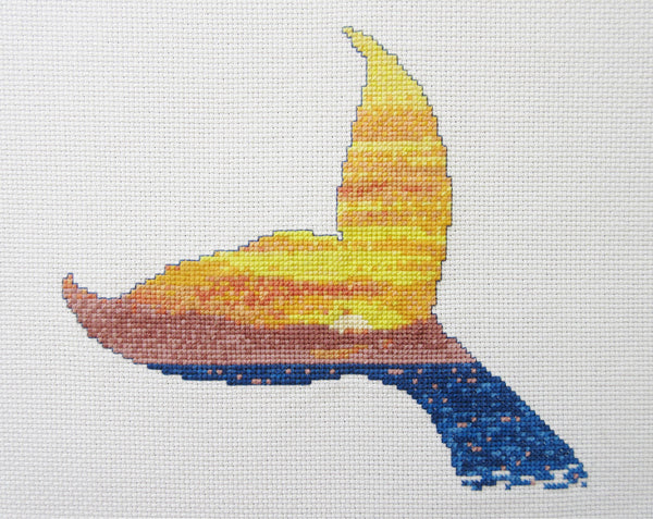 Cross stitch pattern of the silhouette of a whale's tail filled with a scene of a sunset over the ocean. Straight view of stitched piece.