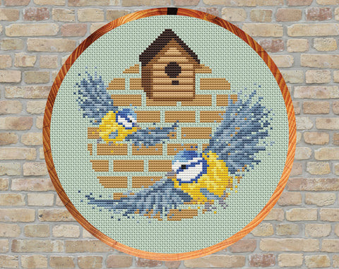 Blue Tits and Birdhouse cross stitch pattern. Two splattered paint style blue tits flying out from a birdhouse. Shown in hoop.