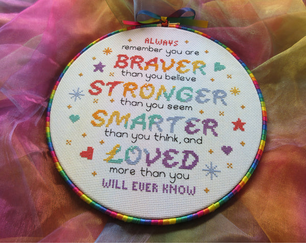 Cross stitch pattern of the quote 'Always remember that you are BRAVER than you believe, STRONGER than you seem, SMARTER than you think, and LOVED more than you will ever know", in rainbow colours. Shown in 10 inch hoop with rainbow ribbon around the hoop, and with the stitched piece at an oblique angle.