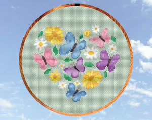 Butterfly Heart cross stitch pattern. Butterflies in pink, purple and blue pastel colours, daisies, yellow flowers and leaves form a heart shape. Shown in hoop.