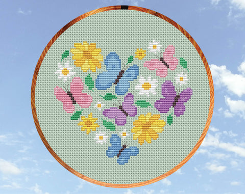 Butterfly Heart cross stitch pattern. Butterflies in pink, purple and blue pastel colours, daisies, yellow flowers and leaves form a heart shape. Shown in hoop.