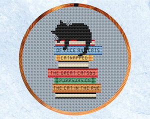 Catnapped cross stitch pattern. Black cat asleep on a pile of books with pun titles. Shown in hoop.