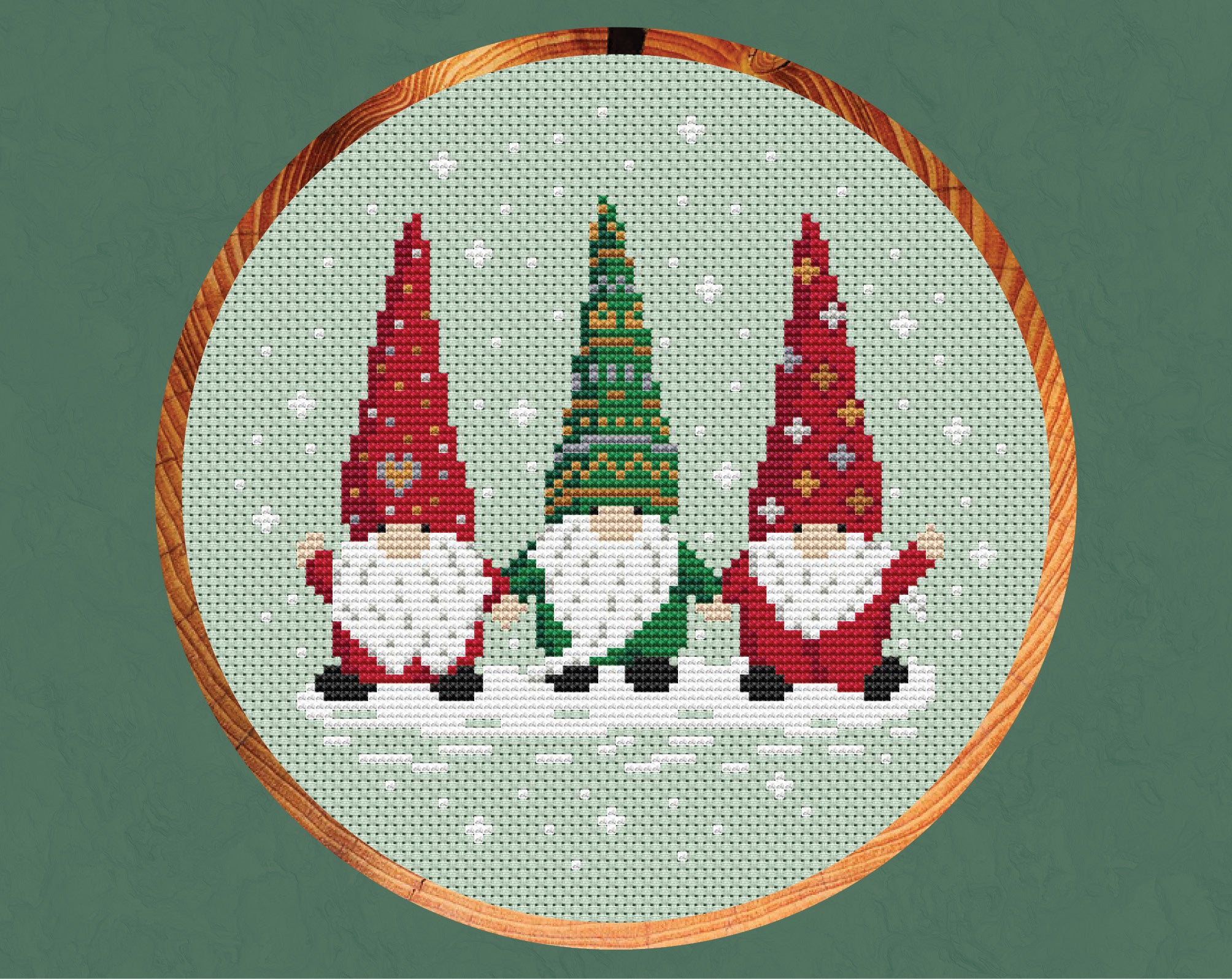 Christmas Gnome Trio cross stitch pattern - three Christmas gonks or nisse holding hands, two in red outfits and one in a green outfit, with a snowy background. Shown in a hoop.