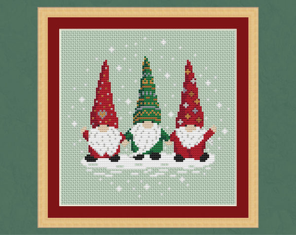 Christmas Gnome Trio cross stitch pattern - three Christmas gonks or tomte holding hands, two in red outfits and one in a green outfit, with white beards and a snowy background. Shown in a frame.