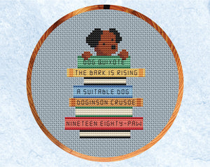 Cross stitch pattern of dog with its paws on a pile of books labelled with dog puns of classic book titles. Shown in hoop.