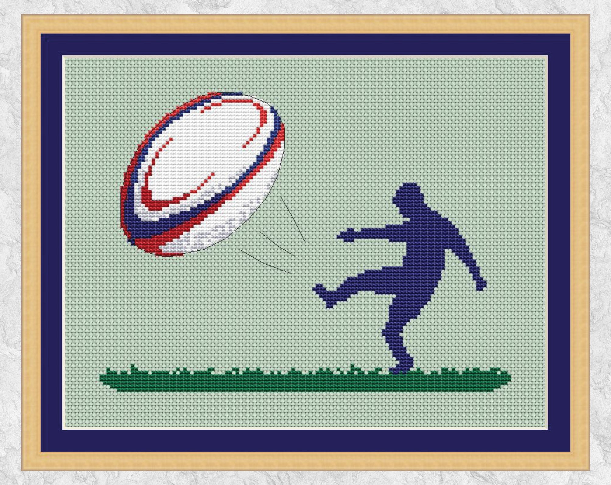 Cross stitch pattern of the silhouette of a rugby player kicking a large colourful rugby ball. Shown with frame.