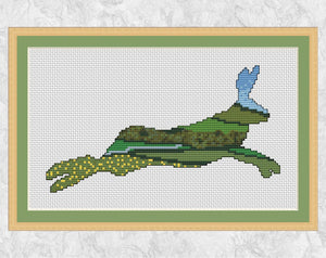 Cross stitch pattern of the silhouette of a running hare, filled with a scene of the British countryside with rolling hills, fields, farmland, buttercups, woodland and a river. Shown with frame.