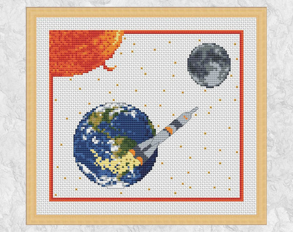 'To the Moon' - Space cross stitch pattern showing rocket, Earth, Sun and Moon. Shown on white fabric with frame.