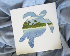 Cross stitch pattern of the silhouette of a turtle filled with a view of a desert island beach, blue sea and sky. Stitched piece displayed on blue fabric background.