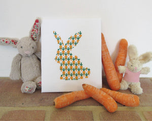 Carrot Bunny cross stitch pattern - silhouette of a rabbit filled with carrots. Stitched piece displayed with toy rabbits and real carrots.