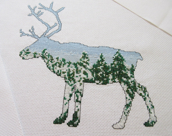 Cross stitch pattern of a reindeer filled with a scene of snow-laden Arctic pine trees against a clear blue sky. Stitched piece.
