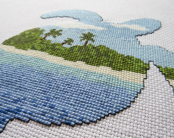 Cross stitch pattern of the silhouette of a turtle filled with a view of a desert island beach, blue sea and sky. Angled view of stitched piece.