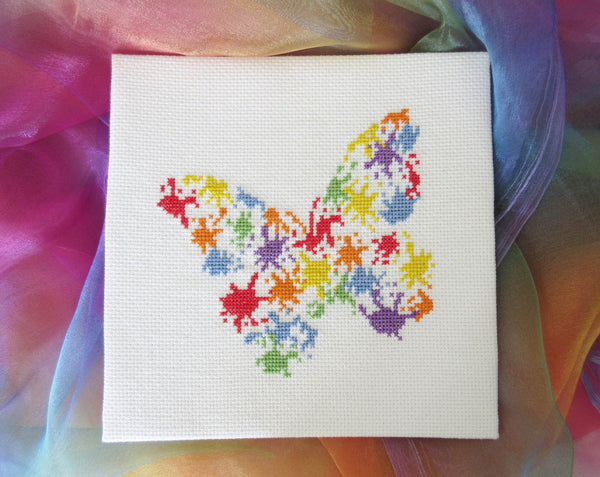 Fun cross stitch pattern of a butterfly made up of splashes of splattered paint. Stitched piece on rainbow background.
