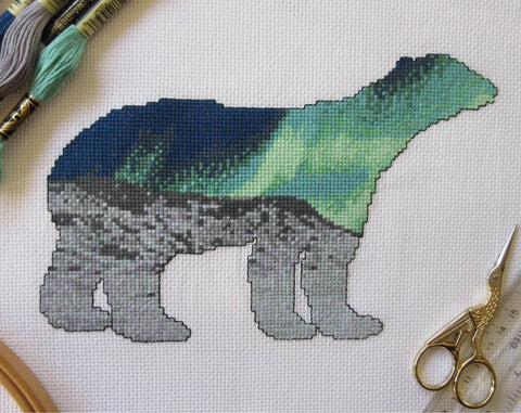 Cross stitch pattern of the silhouette of a polar bear, filled with a scene of the Northern Lights over an Arctic landscape of snow and mountains. Stitched piece with props.