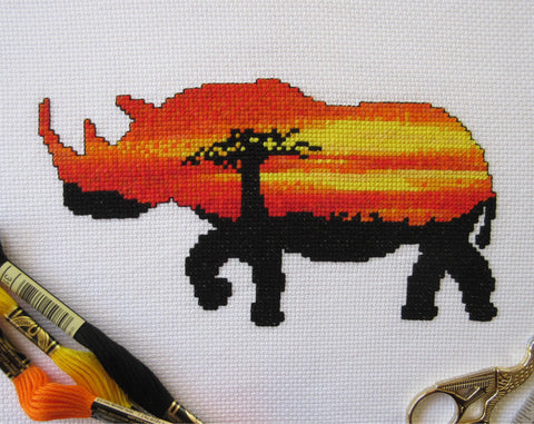 Cross stitch pattern of the silhouette of a rhinoceros filled with a view of an African sunset behind a baobab tree. Stitched piece with props.
