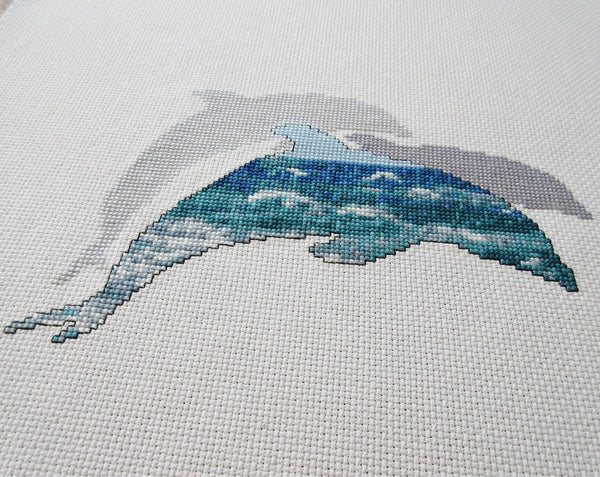 Cross stitch pattern of the silhouette of a dolphin filled with a scene of blue and turquoise ocean waves, with two plain silhouetted dolphins behind it. Angled view of stitched piece.
