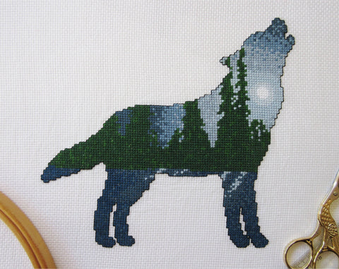 Cross stitch pattern of a wolf silhouette filled with a scene of a moonlit pine forest, with the moonlight reflecting off snow-covered ground. Stitched piece with props.