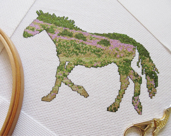Cross stitch pattern of the silhouette of a pony with a view of the New Forest in southern England inside it. Angled view of stitched piece with props.