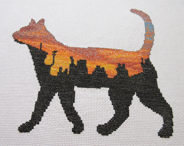 Cross stitch pattern of the silhouette of a cat filled with a scene of a rich and vibrant sunset over city rooftops. Angled view of stitched piece.