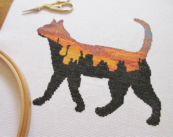 Cross stitch pattern of the silhouette of a cat filled with a scene of a rich and vibrant sunset over city rooftops. Angled view of stitched piece with props.