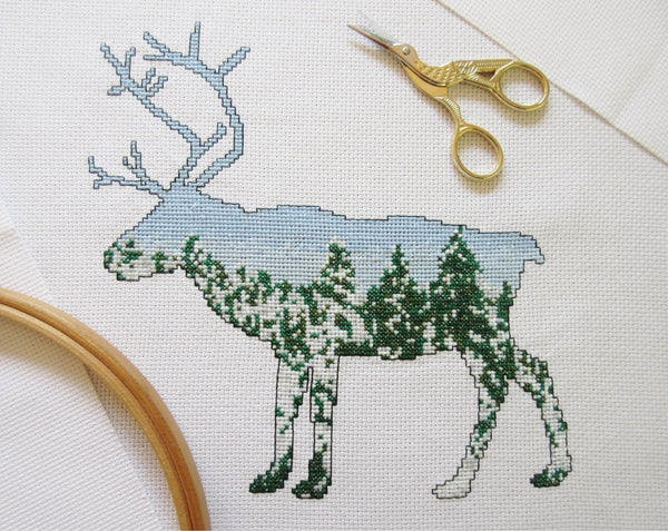 Cross stitch pattern of a reindeer filled with a scene of snow-laden Arctic pine trees against a clear blue sky. Stitched piece with scissors and hoop.