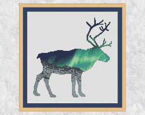 Cross stitch pattern of the silhouette of a reindeer, filled with a scene of the Northern Lights over an Arctic landscape of snow and mountains. Shown with frame.