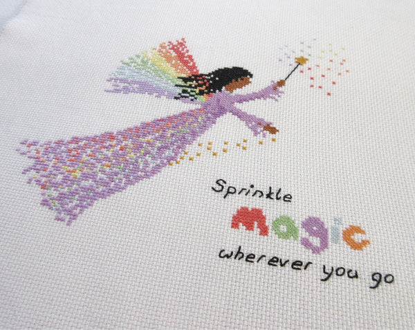 Rainbow Fairy cross stitch pattern - fairy in a purple dress with a wand and wings made of rainbows, and the text 'sprinkle magic wherever you go'. Angled view of stitched piece.