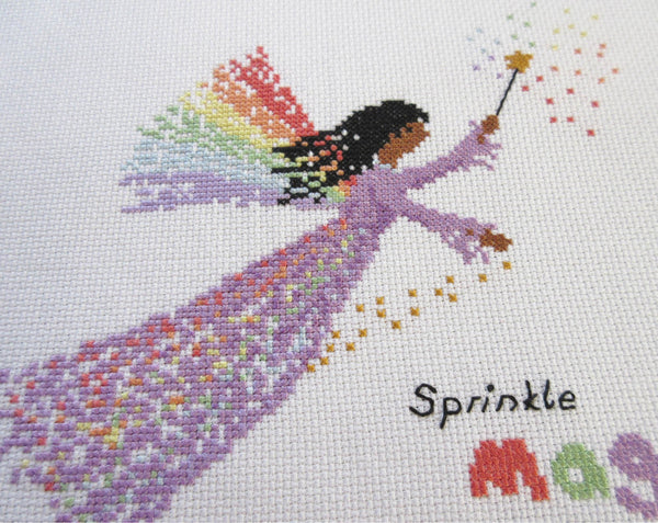 Rainbow Fairy cross stitch pattern - fairy in a purple dress with a wand and wings made of rainbows, and the text 'sprinkle magic wherever you go'. Close up view of stitched piece.