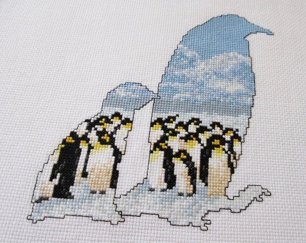 Cross stitch pattern of the silhouette of a penguin and chick, filled with an Antarctic scene of penguins huddled together on the snow, with mountains in the distance below a clear blue sky. Angled view of stitched piece.