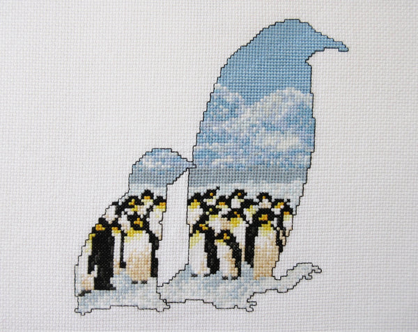 Cross stitch pattern of the silhouette of a penguin and chick, filled with an Antarctic scene of penguins huddled together on the snow, with mountains in the distance below a clear blue sky. Stitched piece without hoop.