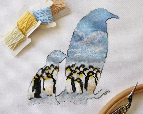 Cross stitch pattern of the silhouette of a penguin and chick, filled with an Antarctic scene of penguins huddled together on the snow, with mountains in the distance below a clear blue sky. Stitched piece with props.