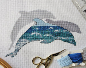 Cross stitch pattern of the silhouette of a dolphin filled with a scene of blue and turquoise ocean waves, with two plain silhouetted dolphins behind it. Stitched piece with props.